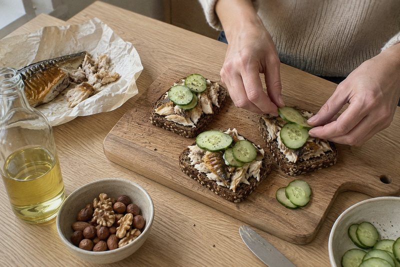Rugbrød med makrel og agurk bliver gjort klar på et skærebræt med en skål nødder ved siden af